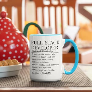 May include: White ceramic mug with a blue interior and handle. The mug features the text "FULL-STACK DEVELOPER" with a definition and the name "Charlotte" at the bottom. The mug is on a brown surface.