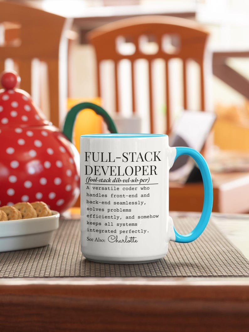 May include: White ceramic mug with a blue interior and handle. The mug features the text "FULL-STACK DEVELOPER" with a definition and the name "Charlotte" at the bottom. The mug is on a brown surface.