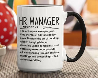 May include: White ceramic mug with a black handle and rim. The mug features the text "HR MANAGER" and a humorous definition, including the name "Sarah". The mug is on a brown surface.