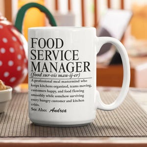 Può includere: Una tazza in ceramica bianca con testo nero che definisce un responsabile del servizio di ristorazione come una mente professionale che organizza le cucine, gestisce i team, soddisfa i clienti e assicura il flusso del cibo. Vedi anche: Andrea.