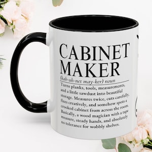 Op de afbeelding: Een witte keramische mok met een zwarte handgreep en zwarte binnenrand. De mok is voorzien van zwarte tekst die de term Cabinet Maker definieert, waarbij het beroep wordt omschreven als iemand die planken en zaagsel omzet in opslagruimte.