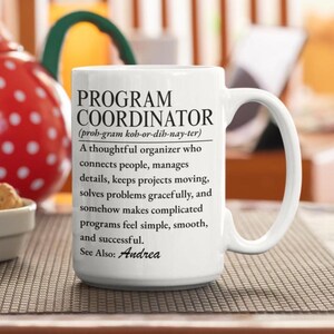 May include: A white ceramic mug featuring black printed text that defines a Program Coordinator as a thoughtful organiser who connects people, manages details, keeps projects moving, solves problems gracefully, and makes programmes successful. The text ends with See Also: Andrea.