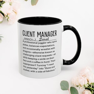 May include: White ceramic mug with a black rim and handle. The mug features a black and white graphic with the text "CLIENT MANAGER noun(n.) Sarah A professional juggler who spins plates, balances expectations, and occasionally wrestles with dragons-otherwise known as challenging client requests-all while keeping a smile on their face and a coffee in hand. Their superpower? Turning "I need this yesterday" into "Done and dusted, with a side of fabulous."
