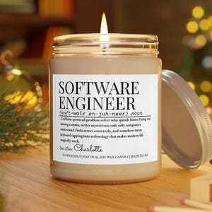 May include: A lit candle in a glass jar with a lid. The label reads "SOFTWARE ENGINEER" with a definition. The candle is on a wooden surface. The text also says "See Also: Charlotte".