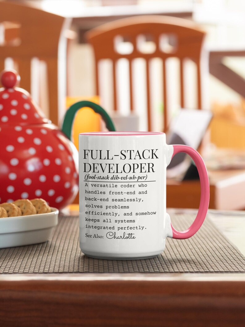 May include: White ceramic mug with a pink interior and handle. The mug features the text "FULL-STACK DEVELOPER" with a definition. The mug is on a patterned surface, with a red and white polka-dotted item in the background.
