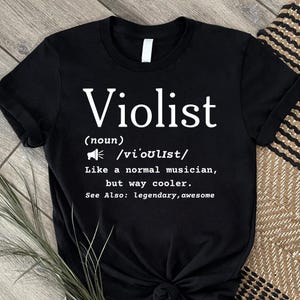 May include: A black short-sleeved t-shirt featuring white text that reads: Violist (noun) /vi'oʊlɪst/ Like a normal musician, but way cooler. See Also: legendary, awesome. The design includes a small megaphone icon next to the phonetic spelling.