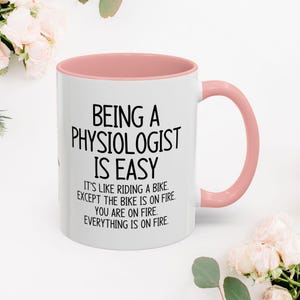 Può includere: Una tazza da caffè in ceramica bianca con un bordo e un manico rosa. La tazza ha un testo nero che dice "Essere un fisiologo è facile. È come andare in bicicletta. Tranne che la bicicletta è in fiamme. Tu sei in fiamme. Tutto è in fiamme."