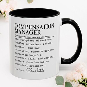 May include: A white ceramic mug with a black interior and handle, featuring black printed text that defines the term Compensation Manager as a workplace wizard who handles salaries, raises, bonuses, and pay questions, ending with See Also: Charlotte.