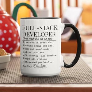 May include: White ceramic mug with a black handle and rim. The mug features the text "FULL-STACK DEVELOPER" and a definition, along with the name "Charlotte". The mug is on a patterned surface.