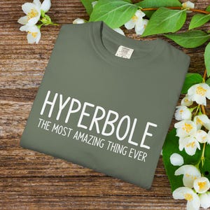 May include: An olive green t-shirt with the word "HYPERBOLE" in large white letters, followed by "THE MOST AMAZING THING EVER" in smaller white letters. The shirt is folded and placed on a wooden surface, decorated with white flowers and green leaves.