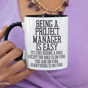 May include: White ceramic coffee mug with a black rim and handle. The mug has black text that reads "Being a Project Manager is Easy. It's like riding a bike. Except the bike is on fire. You are on fire. Everything is on fire."