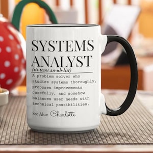May include: A white ceramic mug with a black handle and rim, featuring black text that defines a systems analyst as a problem solver who studies systems, proposes improvements, and balances user needs with technical possibilities, ending with See Also: Charlotte.