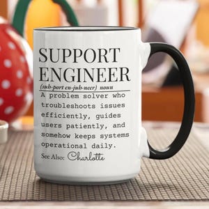 Könnte beinhalten: Ein weißer Keramikbecher mit schwarzem Henkel und Rand, bedruckt mit schwarzem Text, der einen Support-Ingenieur als Problemlöser definiert, der Fehler behebt, Benutzer anleitet und Systeme betriebsbereit hält, ergänzt durch See Also: Charlotte.