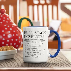 May include: White ceramic mug with a blue handle and rim, featuring the text "FULL-STACK DEVELOPER" and a definition. The mug is on a patterned surface, with a red and white polka-dotted item in the background.