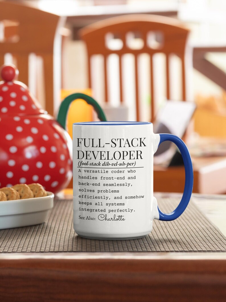 May include: White ceramic mug with a blue handle and rim, featuring the text "FULL-STACK DEVELOPER" and a definition. The mug is on a patterned surface, with a red and white polka-dotted item in the background.