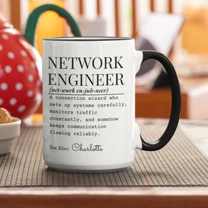 Può includere: Tazza in ceramica bianca con manico e bordo neri. La tazza presenta il testo "NETWORK ENGINEER" con una definizione sottostante. La tazza include anche il nome "Charlotte".