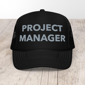 May include: Black trucker hat with the text "PROJECT MANAGER" in gray block letters. The hat has a mesh back and a curved brim. The hat is sitting on a light wood surface.