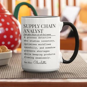 Peut inclure: Mug en céramique blanche avec anse et bord noirs. Le mug affiche le texte "SUPPLY CHAIN ANALYST" et une définition. Le mug est posé sur une surface à motifs.