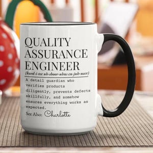 May include: A white ceramic mug with a black handle and rim, featuring black printed text that defines a Quality Assurance Engineer, including a phonetic pronunciation guide and the name Charlotte at the bottom of the descriptive paragraph.