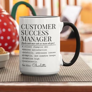 Pode incluir: Caneca de cerâmica branca com alça e borda pretas. A caneca apresenta o texto "CUSTOMER SUCCESS MANAGER" em letras pretas grandes, juntamente com uma definição e o nome "Charlotte".