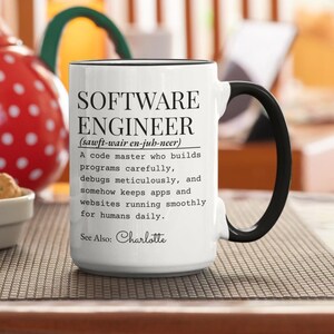 May include: White ceramic mug with black handle and rim. The mug features the text "SOFTWARE ENGINEER" with a phonetic pronunciation and a definition. The text also includes "See Also: Charlotte".