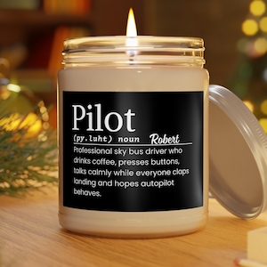 Puede incluir: Una vela encendida en un tarro de cristal con tapa de rosca. El tarro tiene una etiqueta negra con la palabra "Pilot" y una definición humorística. La vela es beige y la llama es amarilla.