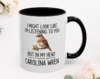 Puede incluir: Taza de cerámica blanca con interior y asa negros. La taza presenta una ilustración de un chochín de Carolina y el texto "I might look like I'm listening to you but in my head I'm thinking about Carolina Wren."