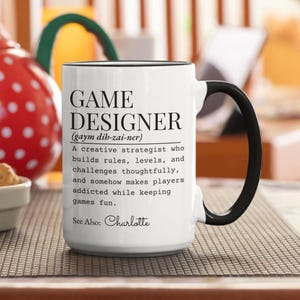 May include: White ceramic mug with a black handle and rim. The mug features the text "GAME DESIGNER" with a definition and the name "Charlotte" at the bottom. The mug is on a brown surface.