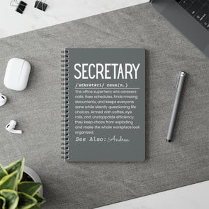 May include: A gray spiral-bound notebook with the word "SECRETARY" in large white letters. The notebook includes a definition of the word and the phrase "See Also: Andrea". A silver pen and other office supplies are also visible.