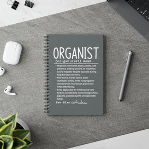 May include: Gray spiral-bound notebook with the word "ORGANIST" in white, along with a definition. The notebook is on a gray surface, with a pen, wireless earbuds, and paper clips nearby. The text includes a definition and related phrases.