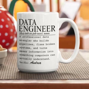May include: A white ceramic mug featuring black printed text that defines a Data Engineer as a professional data wrangler who builds pipelines, fixes broken systems, and turns messy information into something computers can understand. See Also: Andrea.