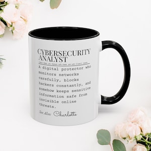 Może przedstawiać: Biały ceramiczny kubek z czarnym wnętrzem i uchwytem. Kubek zawiera tekst "CYBERSECURITY ANALYST" z definicją poniżej. Tekst opisuje cyfrowego obrońcę, który monitoruje sieci i blokuje hakerów. Zawiera również imię "Charlotte".