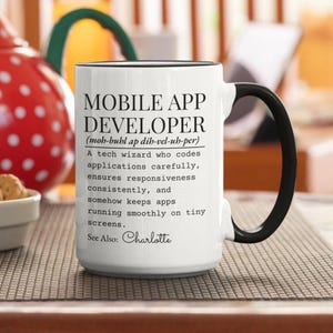 Könnte beinhalten: Weißer Keramikbecher mit schwarzem Henkel und Rand. Der Becher zeigt den Text "MOBILE APP DEVELOPER" mit phonetischer Aussprache und einer Beschreibung der Rolle. Der Name "Charlotte" ist ebenfalls enthalten.