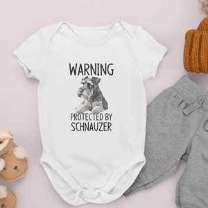 May include: White baby onesie with short sleeves featuring a black and white image of a miniature schnauzer and the text "WARNING PROTECTED BY SCHNAUZER".