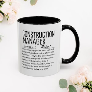 Könnte beinhalten: Weißer Keramikbecher mit schwarzem Innenraum und Henkel. Der Becher zeigt den Text "CONSTRUCTION MANAGER" in fetten schwarzen Buchstaben, zusammen mit einer Definition und dem Namen "Robert". Der Becher ist von Blumen umgeben.