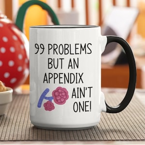May include: White ceramic mug with a black rim and handle. The mug has a black graphic that says "99 problems but an appendix ain't one!" with a cartoon image of an appendix.