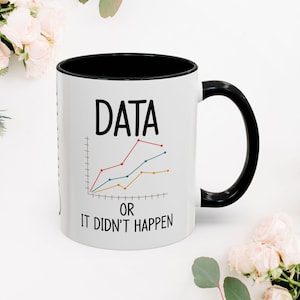 Pode incluir: Caneca de cerâmica branca com interior e alça pretos. A caneca apresenta a palavra "DATA" acima de um gráfico com linhas vermelhas, azuis e amarelas. Abaixo do gráfico, o texto diz "OR IT DIDN'T HAPPEN".