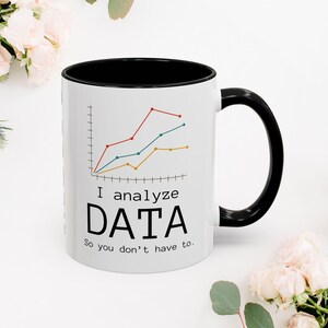 Pode incluir: Caneca de cerâmica branca com interior e alça pretos. A caneca apresenta um gráfico com linhas vermelhas, azuis e amarelas e o texto "I analyze DATA So you don't have to."