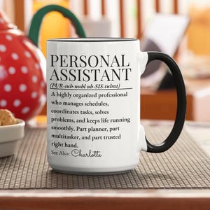 May include: A white ceramic mug with a black handle and rim, featuring black printed text that defines a Personal Assistant. The text includes a phonetic pronunciation, a descriptive paragraph, and the name Charlotte at the bottom.