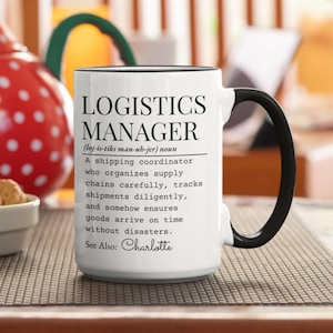 May include: White ceramic mug with a black handle and rim. The mug features the text "LOGISTICS MANAGER" in a bold, black font, along with a definition and the name "Charlotte". The mug is on a patterned surface.