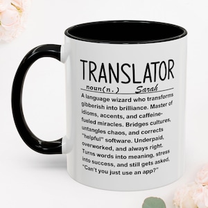 Può includere: Una tazza in ceramica bianca con manico e bordo interno neri. La tazza presenta un testo stampato in nero che definisce la parola traduttore come un mago del linguaggio che trasforma il gergo in brillantezza, incluso il nome Sarah.