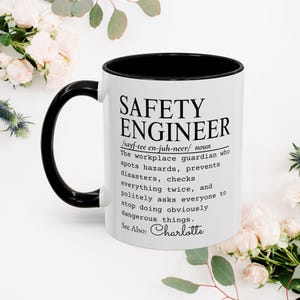 Puede incluir: Una taza de cerámica blanca con interior y asa negros, que presenta un texto en negro que define a un ingeniero de seguridad como un guardián del lugar de trabajo que detecta peligros, evita desastres y pide detener acciones peligrosas.