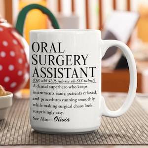 Op de afbeelding: Een witte keramische mok met zwarte bedrukte tekst: ORAL SURGERY ASSISTANT (OR-uhl SUR-juh-ree uh-SIS-tuhnt) A dental superhero who keeps instruments ready, patients relaxed, and procedures running smoothly while making surgical chaos look surprisingly easy. See Also: Olivia