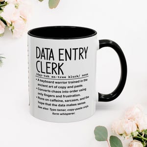 May include: White ceramic mug with a black interior and handle. The mug features the text "DATA ENTRY CLERK" in a bold, black font, along with a humorous definition. The mug is surrounded by white and pink flowers.