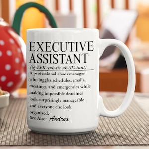 Op de afbeelding: Een witte keramische mok met zwarte bedrukte tekst die een Executive Assistant definieert als een professionele chaosmanager die jongleert met schema's, e-mails, vergaderingen en noodgevallen, terwijl onmogelijke deadlines beheersbaar lijken en iedereen er georganiseerd uitziet. Zie ook: Andrea.