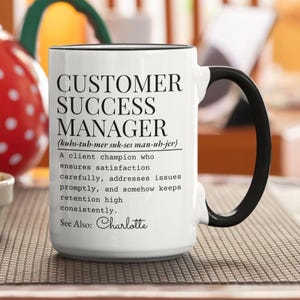 Può includere: Una tazza in ceramica bianca con manico e bordo neri, con testo nero che definisce un Customer Success Manager come un campione del cliente che garantisce soddisfazione, risolve i problemi prontamente e mantiene alta la fidelizzazione, con il nome Charlotte.