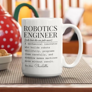 May include: White ceramic mug with the text "ROBOTICS ENGINEER" and a definition. The mug also includes the name "Charlotte". The mug is on a brown surface.