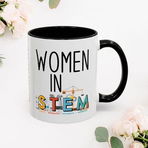 May include: White ceramic mug with a black rim and handle. The mug has the text "Women in STEM" in black and colourful illustrations of science, technology, engineering, and mathematics symbols. The text "Science, Technology, Engineering, Mathematics" is printed in small font below the illustrations.