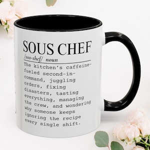May include: A white ceramic mug with a black interior and handle, featuring the black printed text: SOUS CHEF /soo-shef/ noun The kitchen's caffeine-fueled second-in-command, juggling orders, fixing disasters, tasting everything, managing the crew, and wondering why someone keeps ignoring the recipe every single shift.