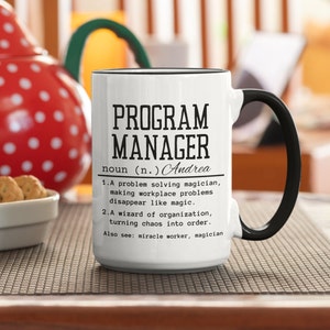 May include: White ceramic coffee mug with a black rim and handle. The mug has black text that reads "PROGRAM MANAGER noun (n.) Andrea 1. A problem solving magician, making workplace problems disappear like magic. 2. A wizard of organization, turning chaos into order. Also see: miracle worker, magician"